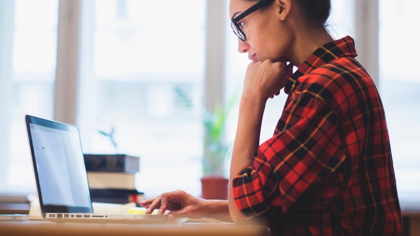 A woman wearing a flannel shirt is sitting at a desk looking at a computer screen.